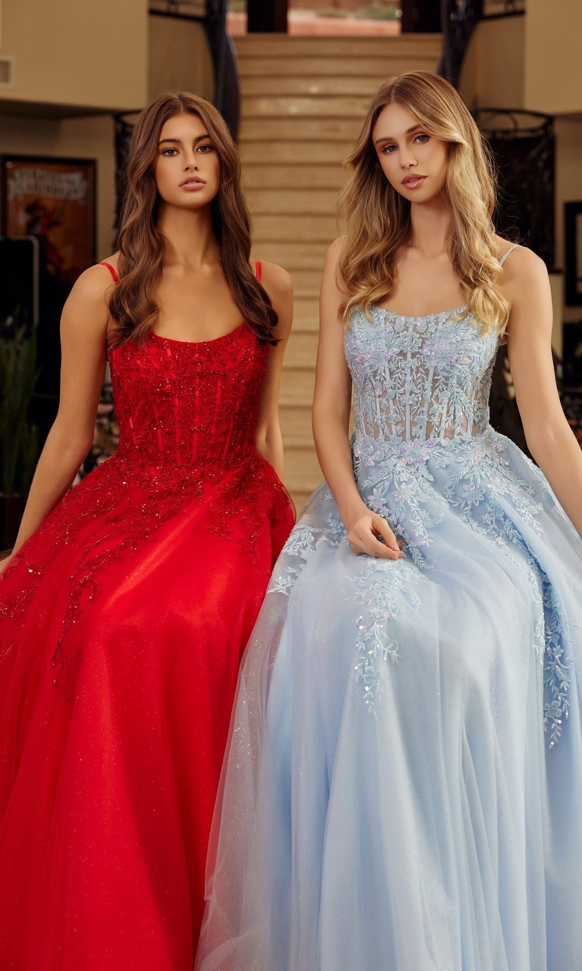 Two women sit together on steps in formal gowns; one is wearing the Nox Anabel H1886 long prom dress, a red sleeveless gown with embellishments, while the other wears a light blue dress with lace and an elegant tulle A-line skirt.